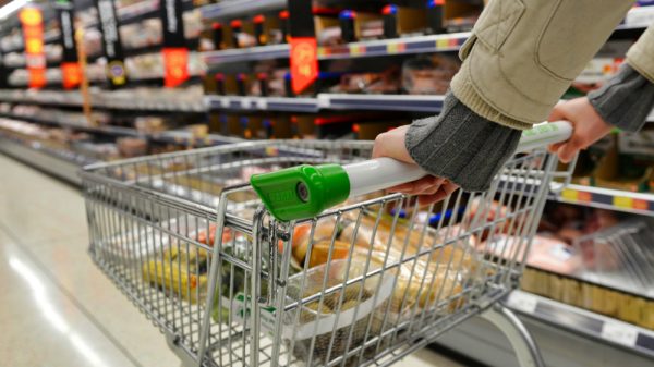A pair of hands push a shopping trolley through a supermarket aisle.