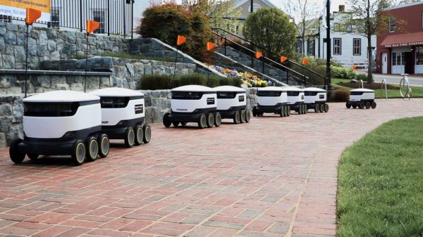 A line of white delivery robots drive down a path.