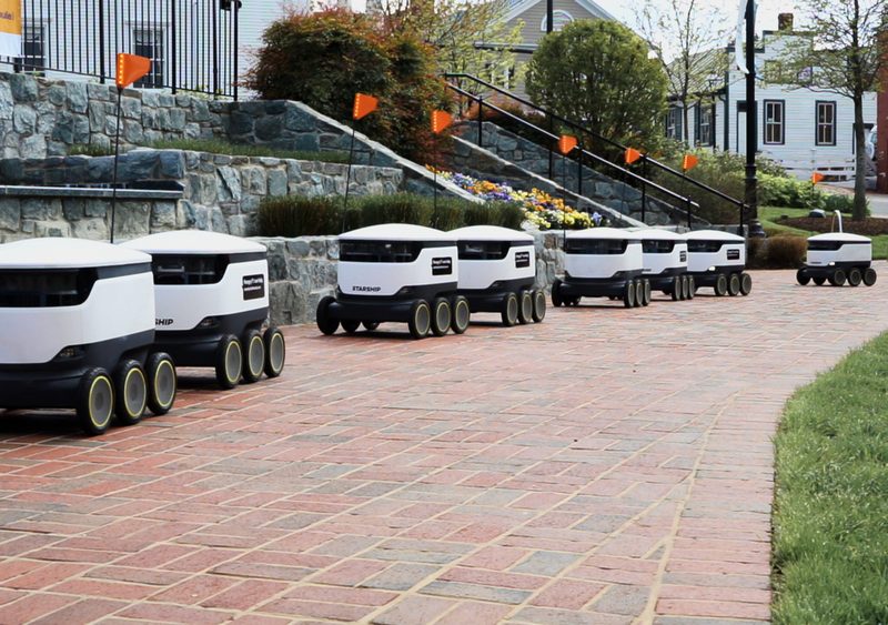 A line of white delivery robots drive down a path.