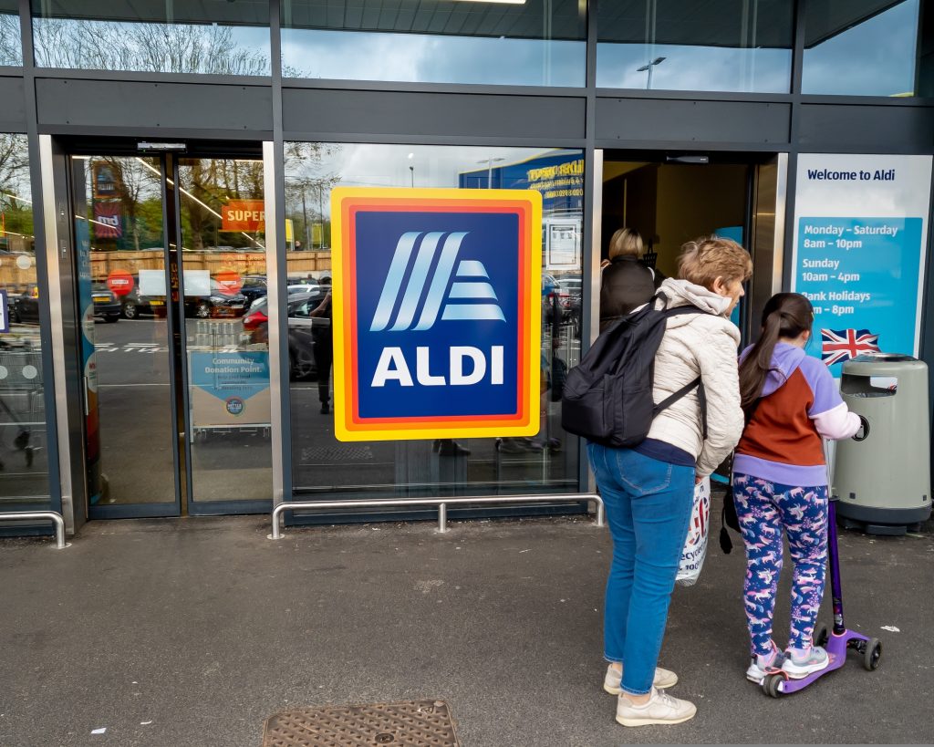 Aldi has revealed another round of price cuts across its range of fresh produce and everyday grocery essentials, here depicting and Aldi store with two customers outside
