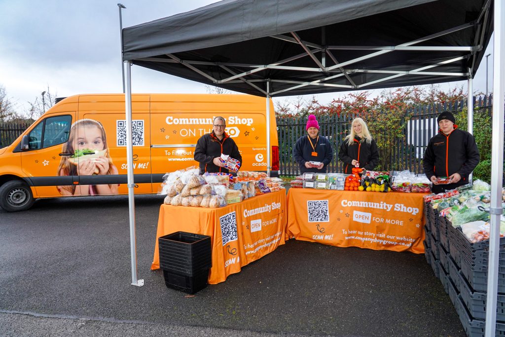 Here showing an Ocado 'On the Go' van in partnership with Community Shop with people in front of an orange table containing food