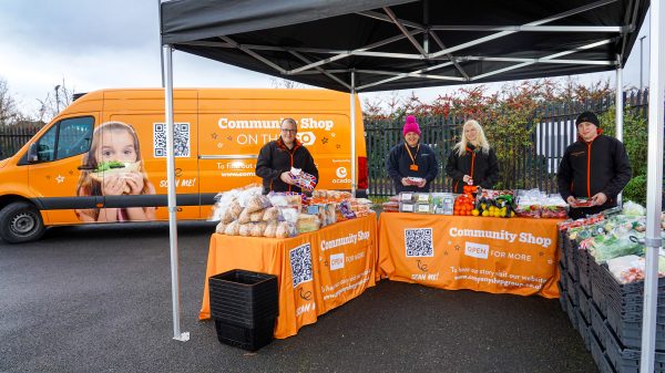 Here showing an Ocado 'On the Go' van in partnership with Community Shop with people in front of an orange table containing food