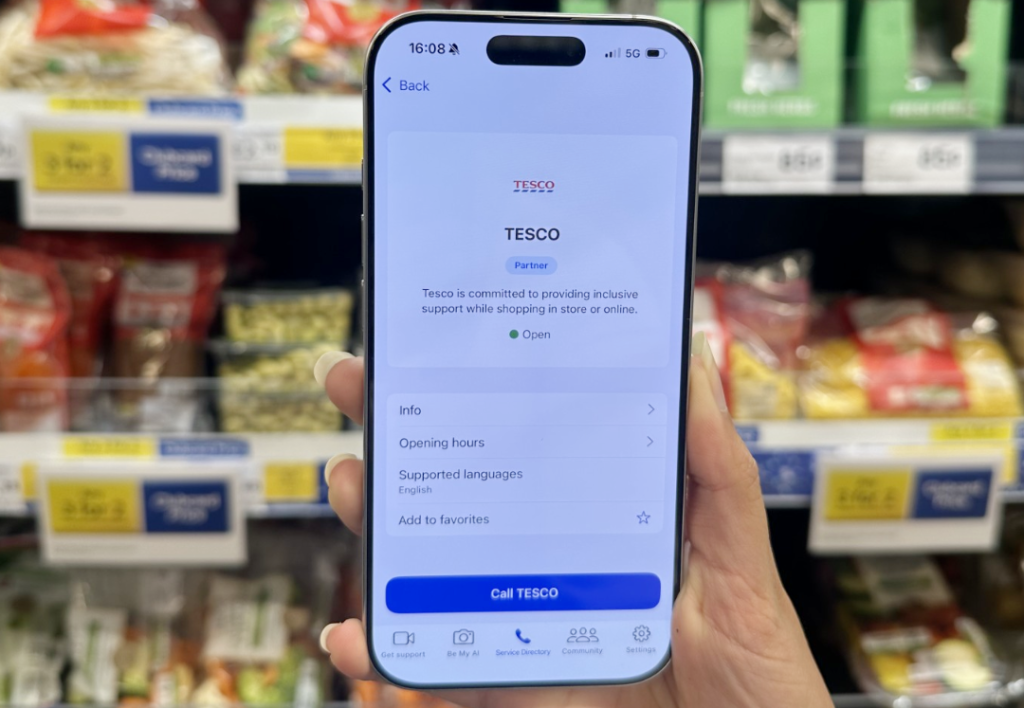 Image depicts an hand holding a smart phone upright in front of Tesco supermarket shelves. On the smart phone, the app 'Be My Eyes' is displayed, as well as 'Tesco' branding.