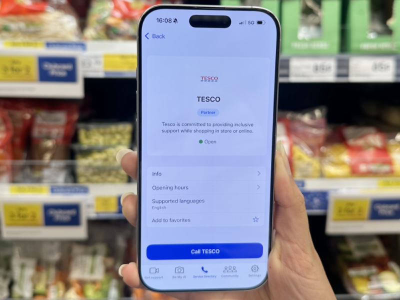 Image depicts an hand holding a smart phone upright in front of Tesco supermarket shelves. On the smart phone, the app 'Be My Eyes' is displayed, as well as 'Tesco' branding.